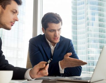 Handsome businessman sitting at desk with partner and pointing on laptop screen when explaining project advantages. Business leaders discussing details of companies collaboration. CEO presenting offer